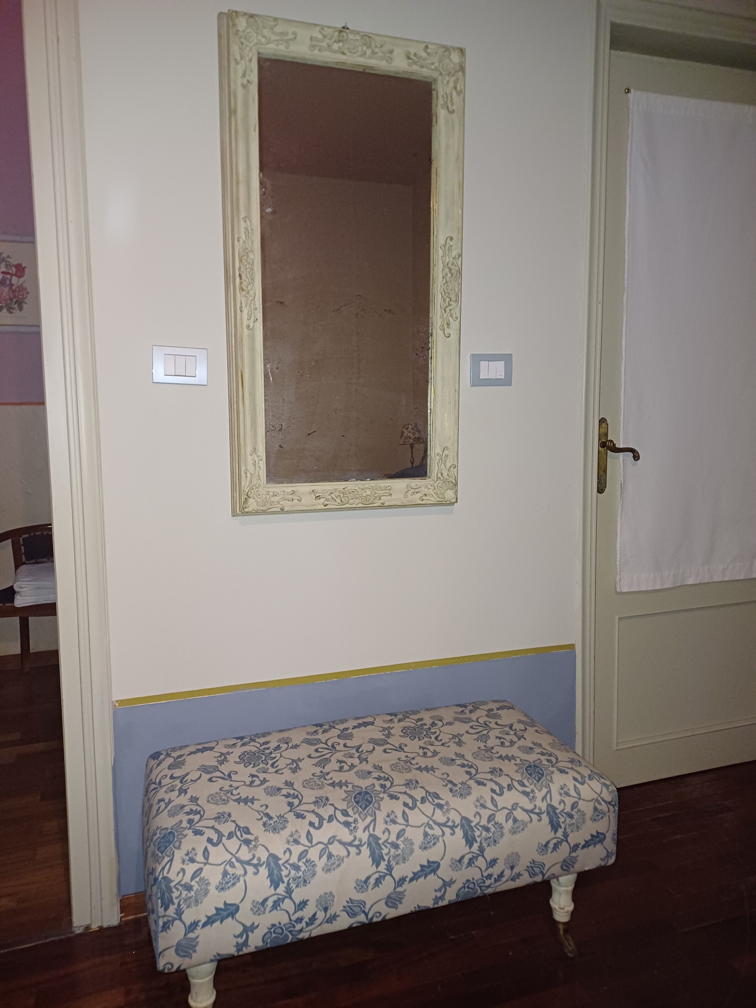 Une pièce avec un mur blanc, un grand miroir doté d’un cadre décoratif, un banc orné d’un motif floral et une porte sur laquelle est accrochée une serviette.