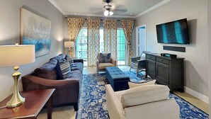 A living room with a sofa, ottoman, and a flat-screen TV. - Coastal Cabana | Beautiful Views + 2 Pools (Galveston)