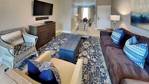 A living room with a television, a sofa, a loveseat, and a coffee table. There is a door leading to another room and a window with curtains. - Coastal Cabana | Beautiful Views + 2 Pools (Galveston)