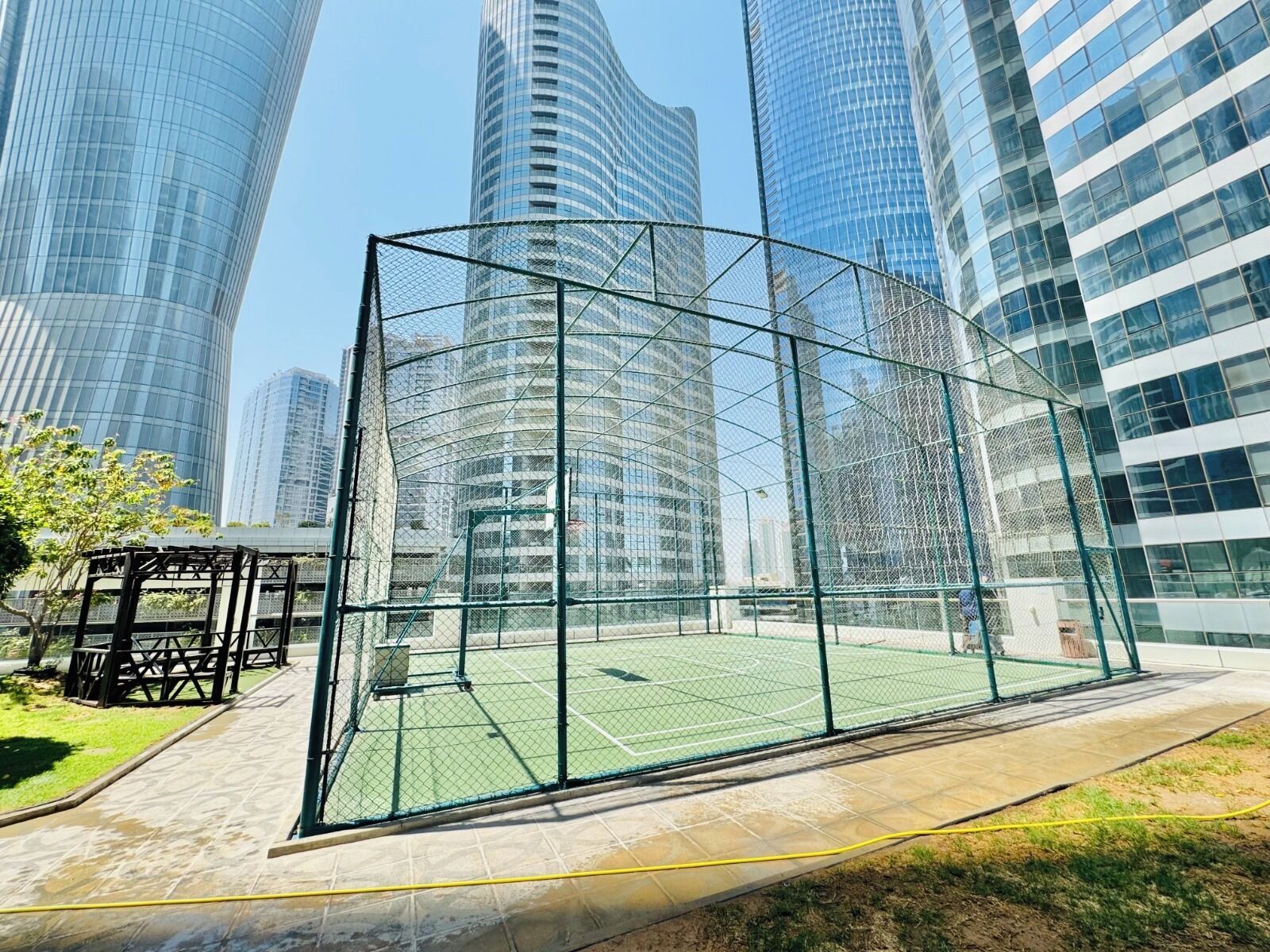 Ein Tennisplatz, umzäunt durch einen Metallzaun, mit einer Laube, umgeben von modernen Hochhäusern.