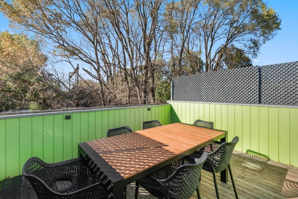 Eine Terrasse im Freien mit einem Holztisch und schwarzen Korbstühlen, umgeben von einem grünen Zaun und Bäumen.
