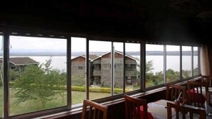 Ein Zimmer mit großen Fenstern und Blick auf einen See und Gebäude. Es gibt Holzstühle und einen Tisch.