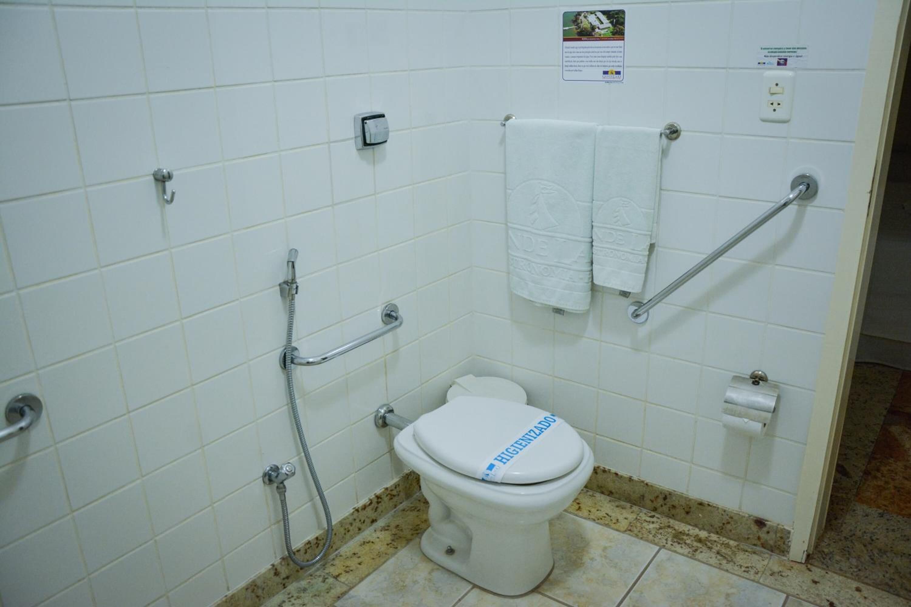Egy fürdőszoba, amelyben fehér WC-papír-tartó, kapaszkodók, törölközőtartó és kézszárító található.