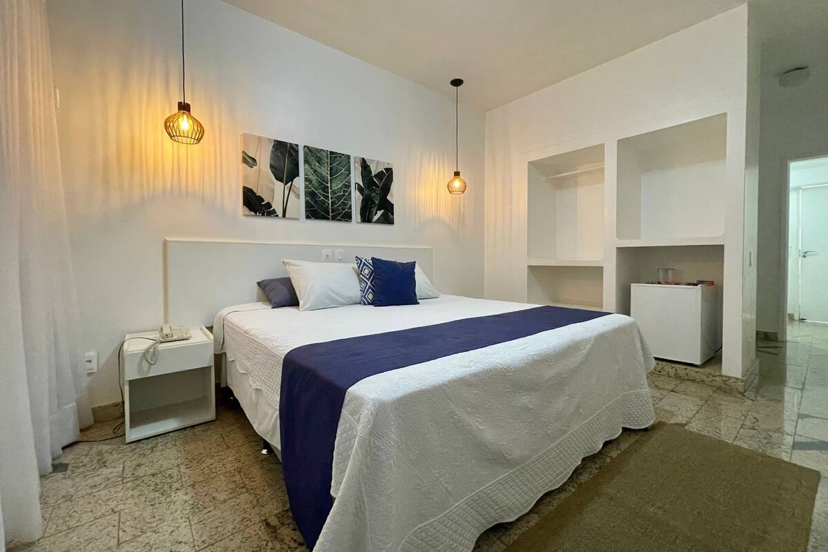 Un dormitorio con cama, mesitas de noche, lámpara de pared y refrigerador.