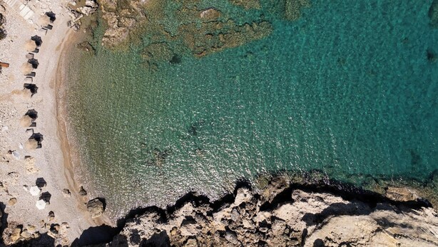 An aerial view of a beach with clear turquoise water, rocky terrain, and scattered umbrellas. - Laftira's Sea View Home (Stegna)