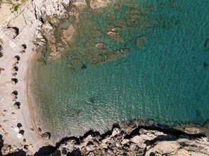 An aerial view of a beach with clear turquoise water, rocky terrain, and scattered umbrellas.