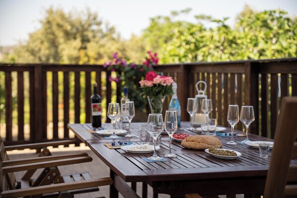 An outdoor wooden table set for a meal with wine glasses, plates, and a loaf of bread.