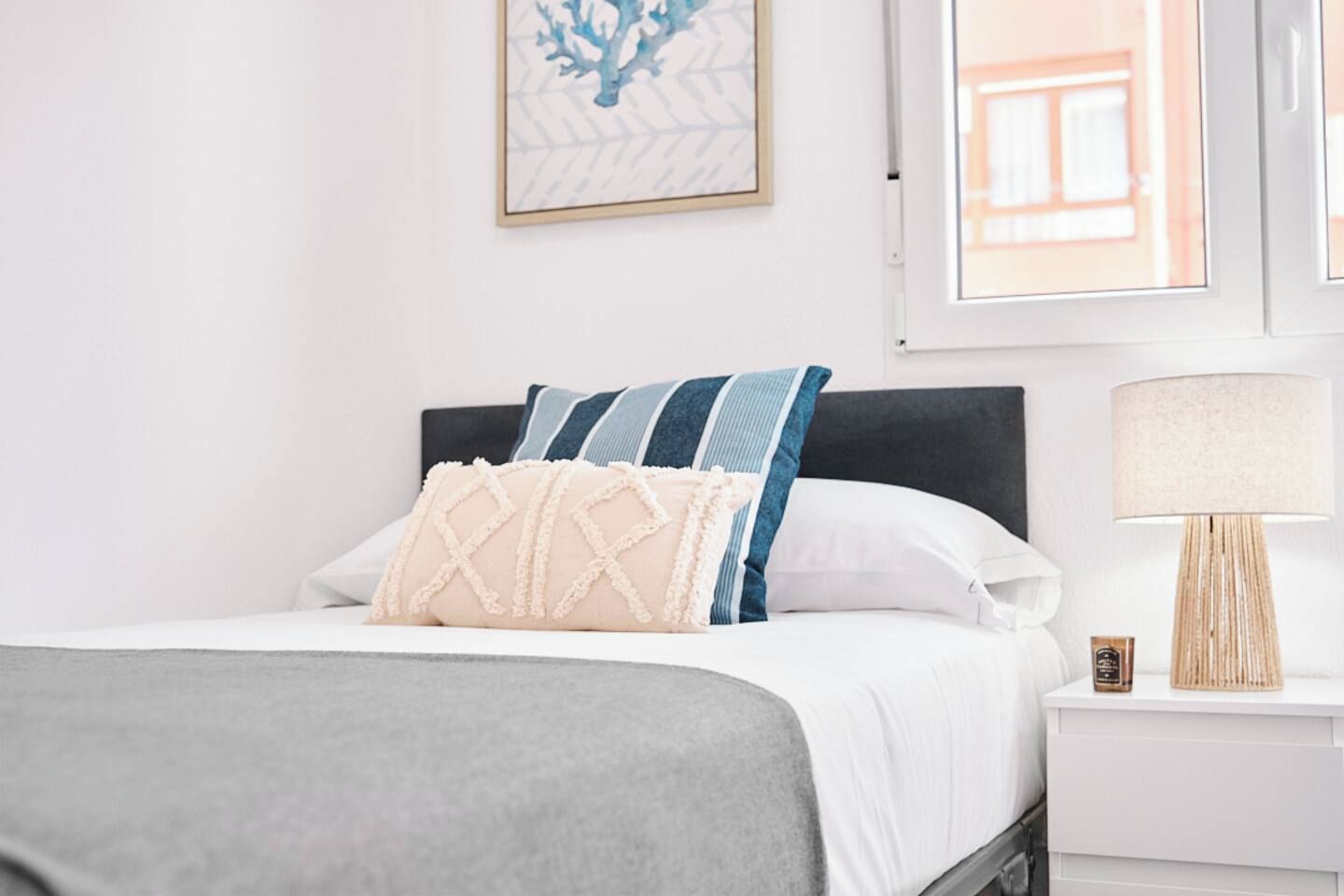 Un lit bien fait, recouvert d’une couvre-lit grise, avec un oreiller blanc et un autre oreiller à rayures bleues et blanches. Au-dessus du lit, on aperçoit une œuvre d’art encadrée, et une lampe est posée sur la table de chevet à côté du lit.