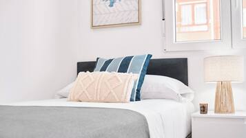 Een net opgemaakt bed met een grijze dekbedovertrek, een wit kussen en een kussen met blauwe en witte strepen. Daarboven hangt een lijst met een kunstwerk en op de nachtkastje staat een lamp.