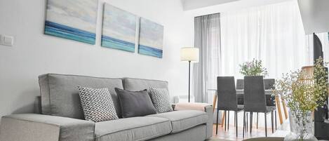 Uma sala de estar moderna com um sofá cinzento, almofadas estampadas, uma mesa de jantar com cadeiras e um vaso com flores.