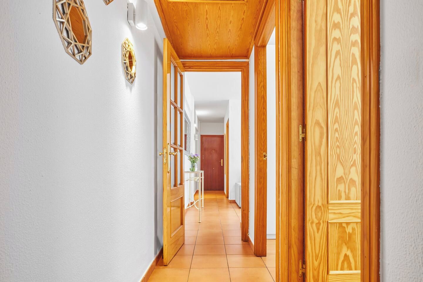 Un pasillo con puertas de madera y suelo de baldosas, que conduce a una habitación con una cama y una mesita de noche.