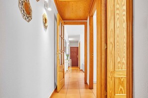 Un couloir avec des portes en bois et un sol carrelé, qui mène à une pièce contenant un lit et une table de chevet.
