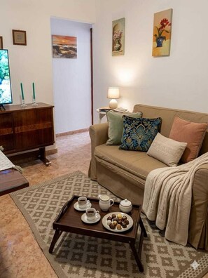 Una sala de estar con un sofá, una mesa de centro con bebidas y aperitivos, un televisor y obras de arte en las paredes.