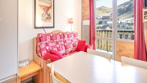 A cozy room with a patterned sofa, a dining table, and a large window offering a view of a town and hills. - Studio in Chamrousse Near Ski Resort (Chamrousse)