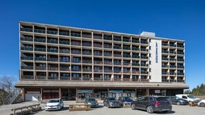 A multi-story hotel with a central entrance, several ground-level shops, and a parking area with cars. - Studio in Chamrousse Near Ski Resort (Chamrousse)