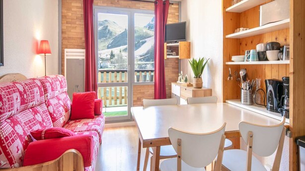 A cozy living area with a sofa, a dining table, and chairs. There is a TV mounted on the wall, a window with a view of mountains, and a kitchen area with shelves and appliances. - Studio in Chamrousse Near Ski Resort (Chamrousse)