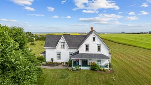 Una casa blanca de dos pisos con techo oscuro, rodeada de campos verdes y un árbol.