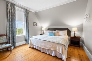 Un dormitorio con una cama grande, suelo de madera, una ventana con cortinas, una mesita de noche con lámpara y una silla.