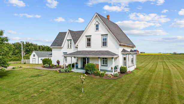 Een groot wit huis met een veranda, omgeven door een verzorgd gazon en een veld op de achtergrond.