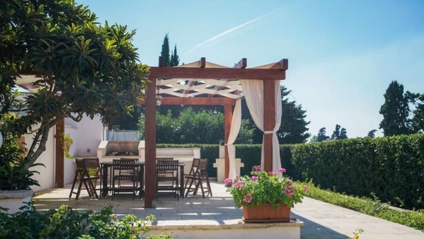 Uma área de estar coberta ao ar livre com pérgola, churrasqueira e um vaso com flores cor-de-rosa.