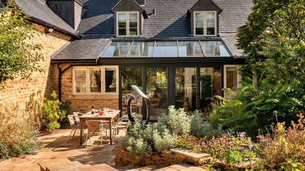 Ein Haus mit Wintergarten, Steinterrasse und einem Garten mit verschiedenen Pflanzen.