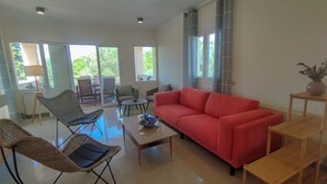 A spacious living room with a red sofa, wicker chairs, a coffee table, and a view of the outdoors through large windows. - Villa Yakinthi (CORFU)