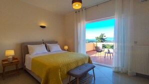 A hotel room with a bed, bedside lamps, a bench, and a view of a balcony with a chair and table. - Villa Yakinthi (CORFU)