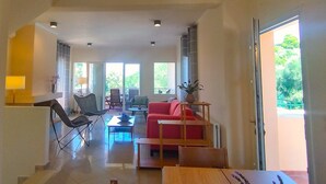 A spacious living area with a red sofa, a wooden coffee table, and a dining table. There are multiple chairs and a lamp. The room has large windows allowing natural light and views of greenery outside. - Villa Yakinthi (CORFU)