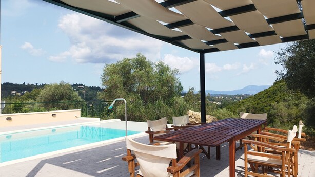 An outdoor pool area with a wooden table and chairs, a pergola overhead, and a view of a hilly landscape. - Villa Yakinthi (CORFU)