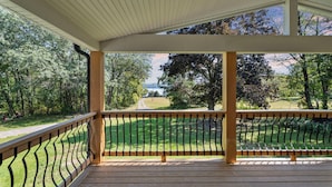 A covered outdoor area with a wooden deck and metal railing, overlooking a grassy landscape with trees and a path.