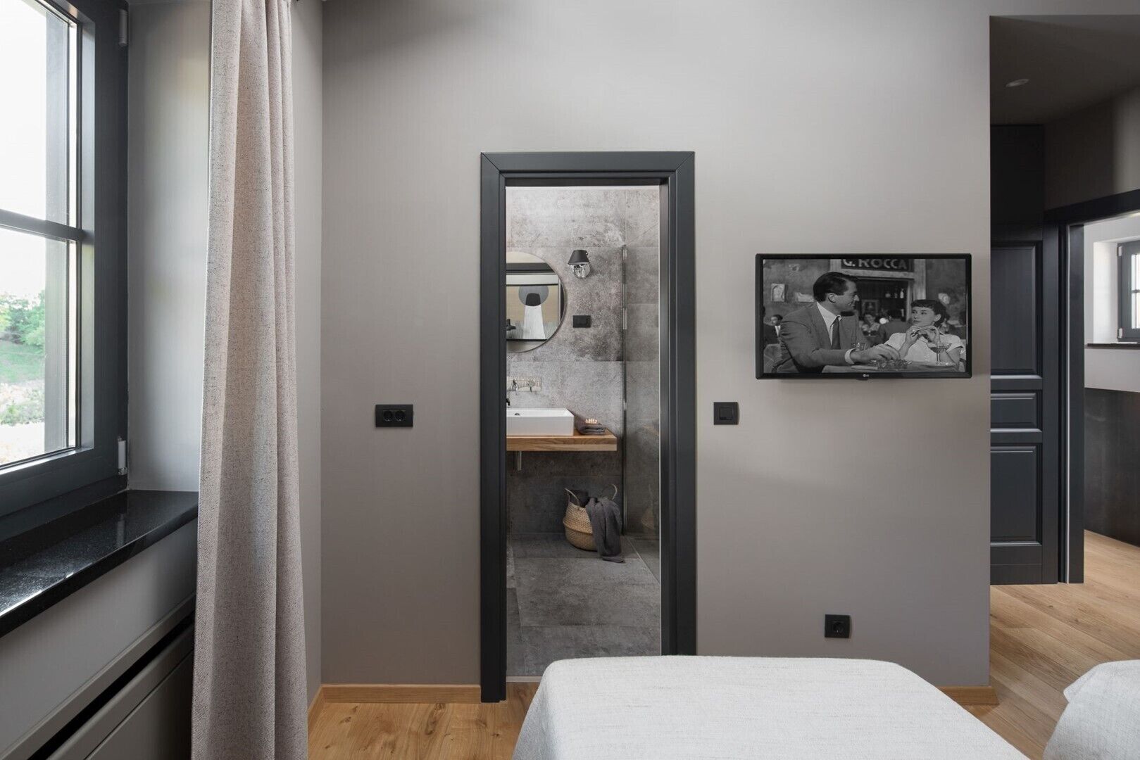 A modern hotel room with a wooden floor, a large window with curtains, a bathroom with a mirror and sink, and a framed picture on the wall.