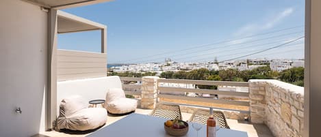 A balcony with a white table, a cushioned chair, and a wicker basket with fruits and a bottle.