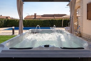 Un jacuzzi con dos grifos, rodeado por una zona cubierta con cortinas, una piscina y un seto.