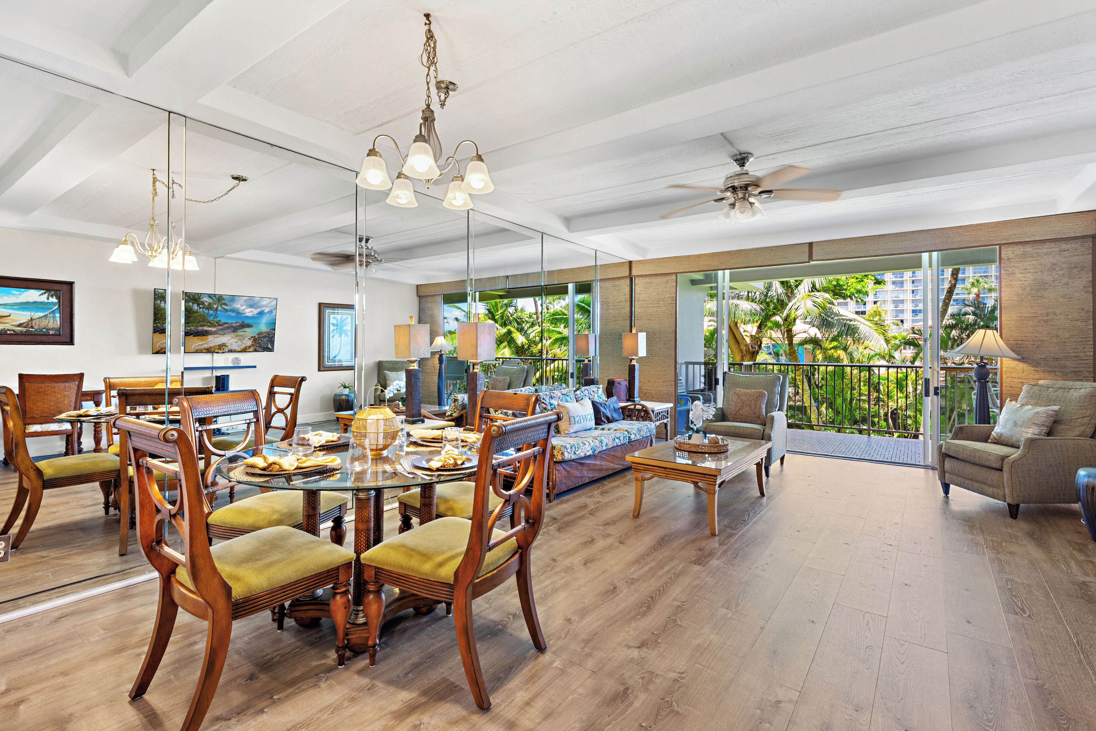 A spacious dining area with wooden chairs and a glass table, a living room with a sofa and chairs, and a balcony with a view of palm trees.