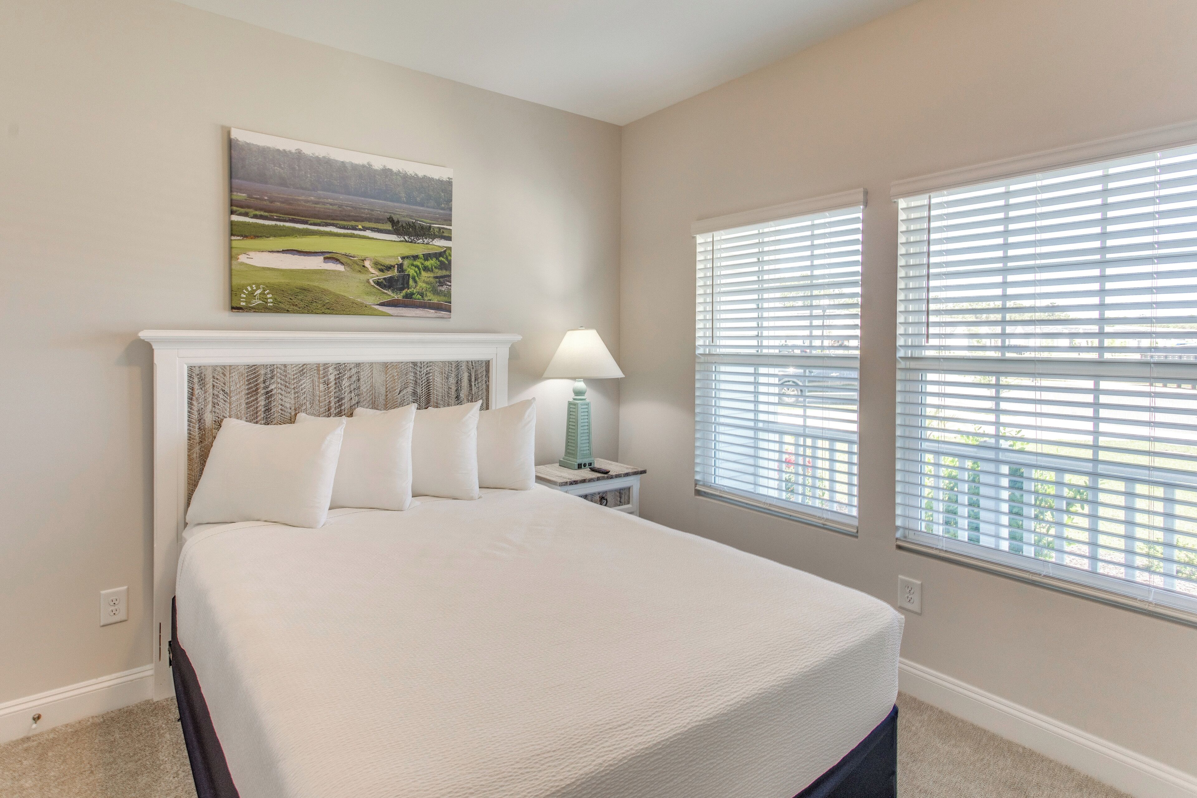 A bedroom with a large bed, a nightstand with a lamp, a painting on the wall, and a window with blinds.