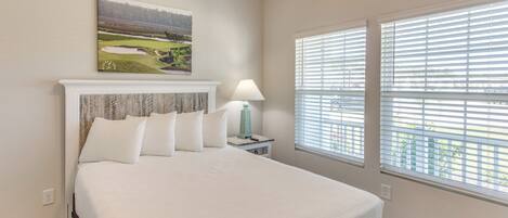 A bedroom with a large bed, a nightstand with a lamp, a painting on the wall, and a window with blinds.