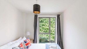 A bedroom with a bed, pillows, a lamp, and a window with curtains. - GuestReady - Spacious 1BR in East London w Balcony (London)