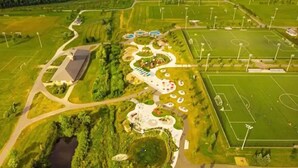 Vista aérea de un parque con un parque infantil, campos deportivos y un edificio de techo inclinado.