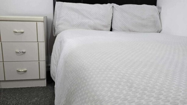 Een bed met een wit beddekleed met patroon en twee kussens, een nachtkastje met lades en een zwart hoofdbord.