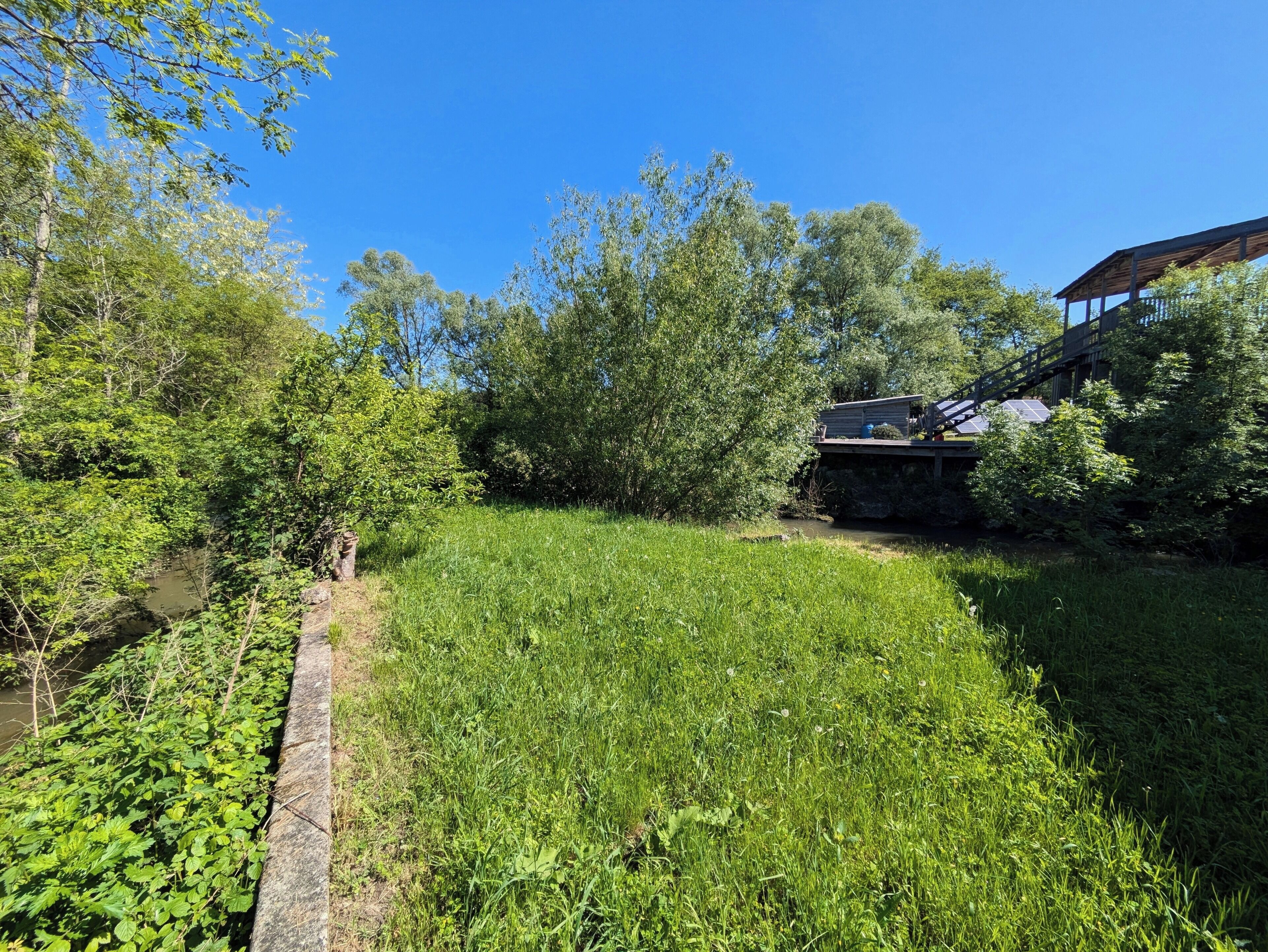 A rustic cabin with a wooden deck and solar panels, surrounded by lush greenery and a small stream.