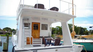 A boat with a covered deck area, a small table with chairs, a wooden door, and a ladder leading to an upper deck.
