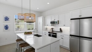 A modern kitchen with white cabinetry, a stainless steel refrigerator, a microwave, and a large island with a sink. - Ocean Pearl - Steps from the Beach (Fort Myers Beach)