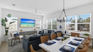 A modern living room with a sofa set, dining table, and large windows with blinds. - Ocean Pearl - Steps from the Beach (Fort Myers Beach)
