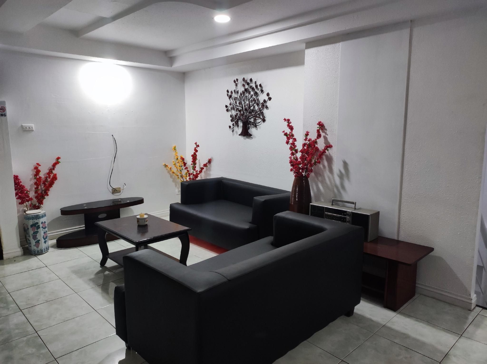 A living room with a black sofa, a coffee table, and two side tables. There are decorative plants and a wall-mounted art piece.