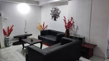 A living room with a black sofa, a coffee table, and two side tables. There are decorative plants and a wall-mounted art piece.
