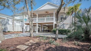 A two-story house with a balcony, a covered porch, and a small garden area with palm trees and shrubs. - Sandy Toes Bungalow: Dog-Friendly with Private Pool (Oak Island)