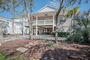 A two-story house with a balcony, a covered porch, and a small garden area with palm trees and shrubs.