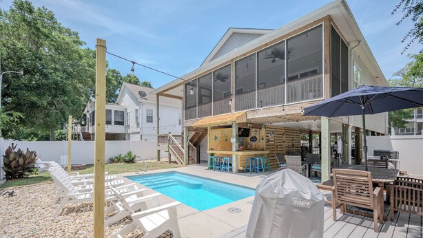 A backyard with a swimming pool, wooden deck, outdoor seating, and a covered grill area. - Sandy Toes Bungalow: Dog-Friendly with Private Pool (Oak Island)