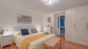 Et soveværelse med seng, sengeborde, bænk, garderobe og et badeværelse, der kan ses igennem en åben dør.