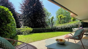 Un espace patio avec une table ronde et deux chaises en osier, un saladier de fruits sur la table, et une vue sur un jardin bien entretenu, agrémenté de divers arbres et arbustes.